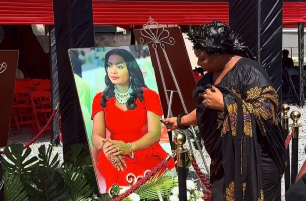 Beverly | Ghanaguardian.com Person in black ceremonial attire gestures toward a large portrait of a woman in a red dress displayed on an easel at an event.