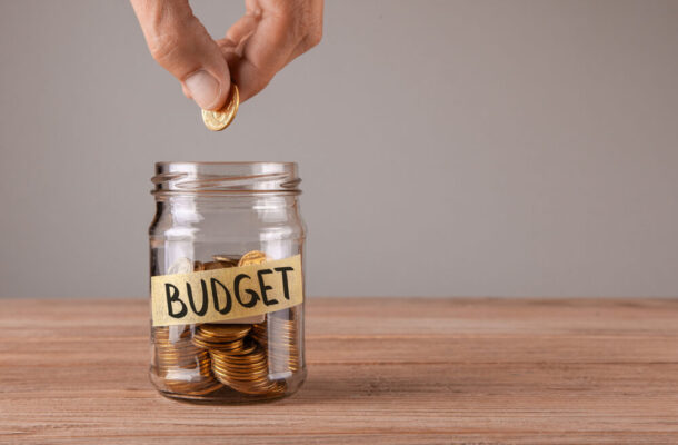 Person drops a coin into a glass jar labeled 'BUDGET' with coins inside on a wooden table, illustrating saving.