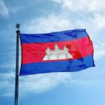 Cambodian national flag waving on a flagpole with Angkor Wat centerpiece against a blue sky.