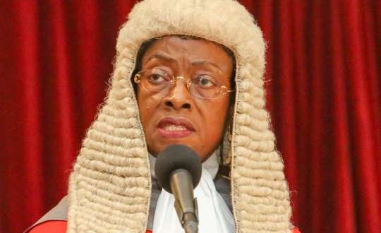 Judge wearing a traditional powdered wig speaking into a microphone on a stage with a red curtain behind them.