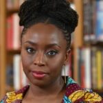Portrait of a Black woman with braided hair wearing a colorful patterned top in a library setting, looking at the camera.