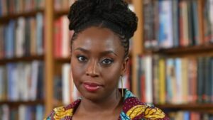 Portrait of a Black woman with braided hair wearing a colorful patterned top in a library setting, looking at the camera.