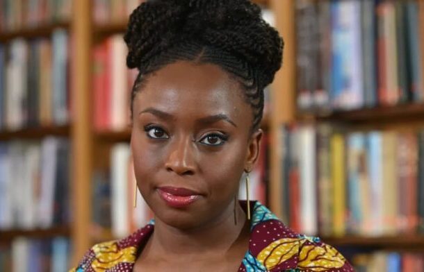 Portrait of a Black woman with braided hair wearing a colorful patterned top in a library setting, looking at the camera.