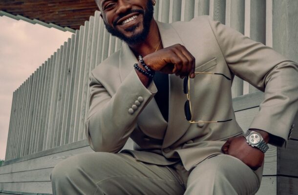 Smiling man in a light beige suit sitting on wooden steps, holding sunglasses in front of him.