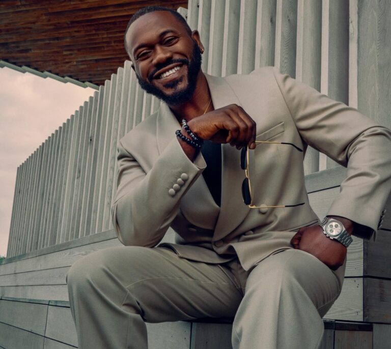 Smiling man in a light beige suit sitting on wooden steps, holding sunglasses in front of him.