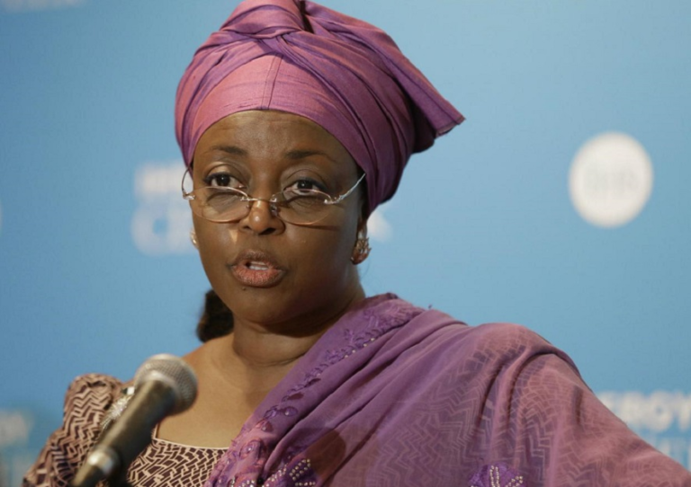 Woman wearing a purple head wrap and glasses speaks into a microphone at a press event.