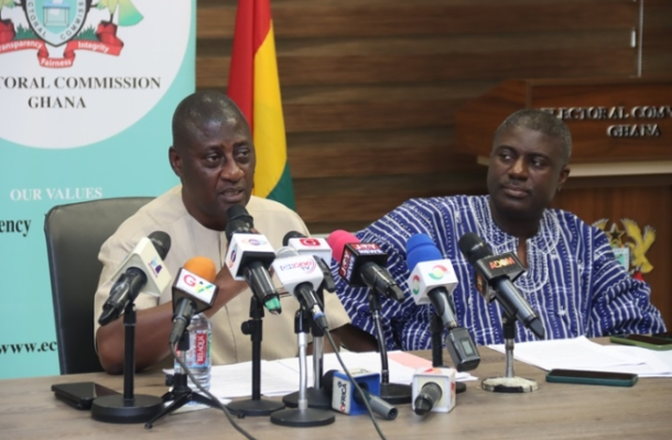Two men sit at a table during a press conference, microphones in front, with a Ghana flag and Electoral Commission banner behind them.