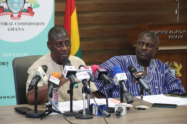 Two men sit at a table during a press conference, microphones in front, with a Ghana flag and Electoral Commission banner behind them.