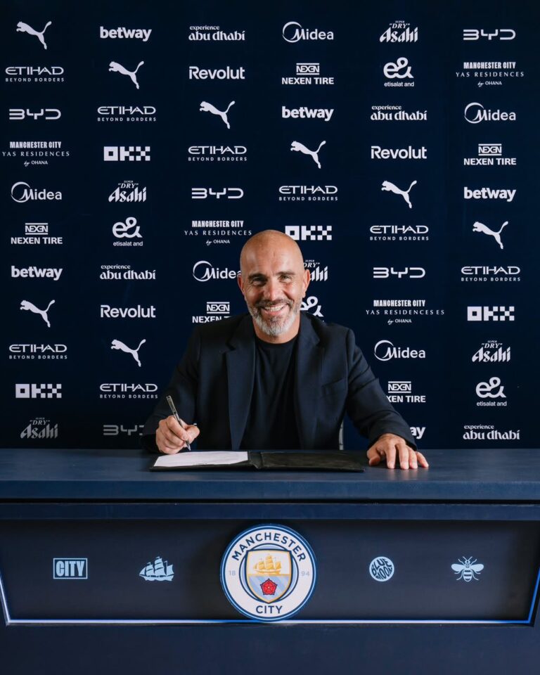 Man in a dark blazer signs a document at a desk, Manchester City logos and sponsor backdrop behind him