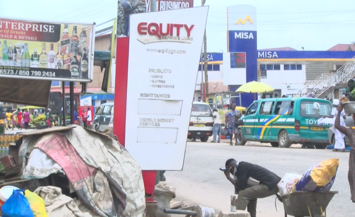 Street scene with an Equity sign advertising loans and savings; market stalls and a green-blue minivan in the background.