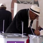 Man in a white hat casts a ballot into a ballot box at a polling station, with others nearby.