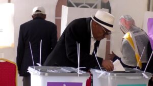 Man in a white hat casts a ballot into a ballot box at a polling station, with others nearby.