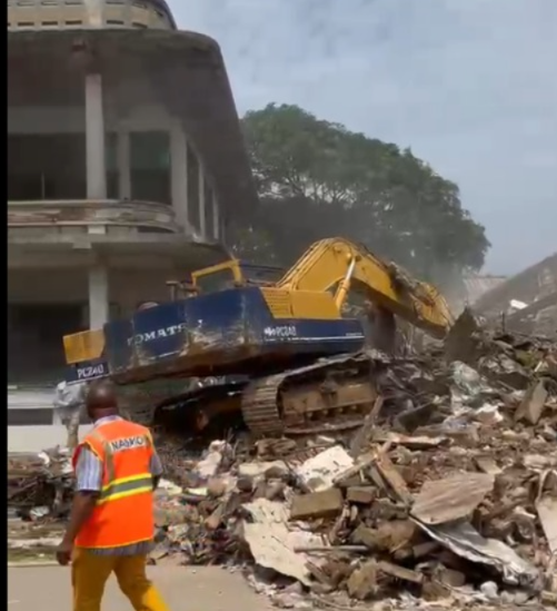 Excavator on a pile of rubble demolishing a building, with a worker in a bright orange safety vest nearby.