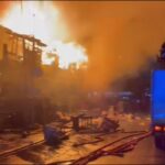 Fire rages on a city street as flames engulf buildings; firefighters stand on a blue fire truck fighting the blaze amid heavy smoke.