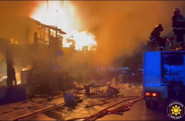 Fire rages on a city street as flames engulf buildings; firefighters stand on a blue fire truck fighting the blaze amid heavy smoke.
