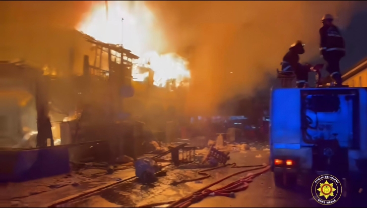 Fire rages on a city street as flames engulf buildings; firefighters stand on a blue fire truck fighting the blaze amid heavy smoke.