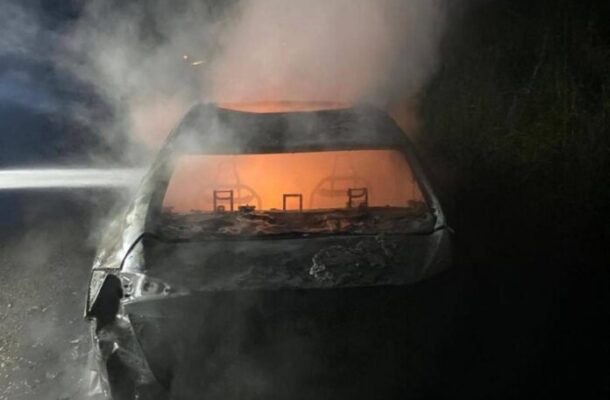 Rear view of a charred car on a dark road at night, orange glow inside and heavy smoke rising above the vehicle.