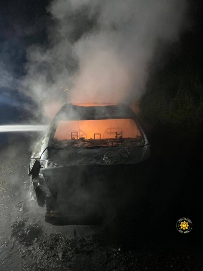 Rear view of a charred car on a dark road at night, orange glow inside and heavy smoke rising above the vehicle.
