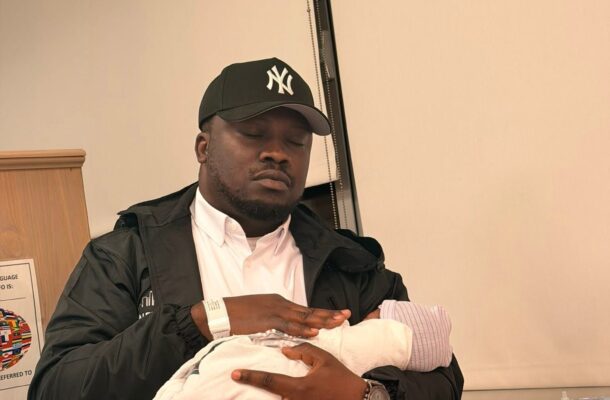Man wearing a NY Yankees cap tenderly holding a newborn swaddled in a white blanket in a clinic setting.