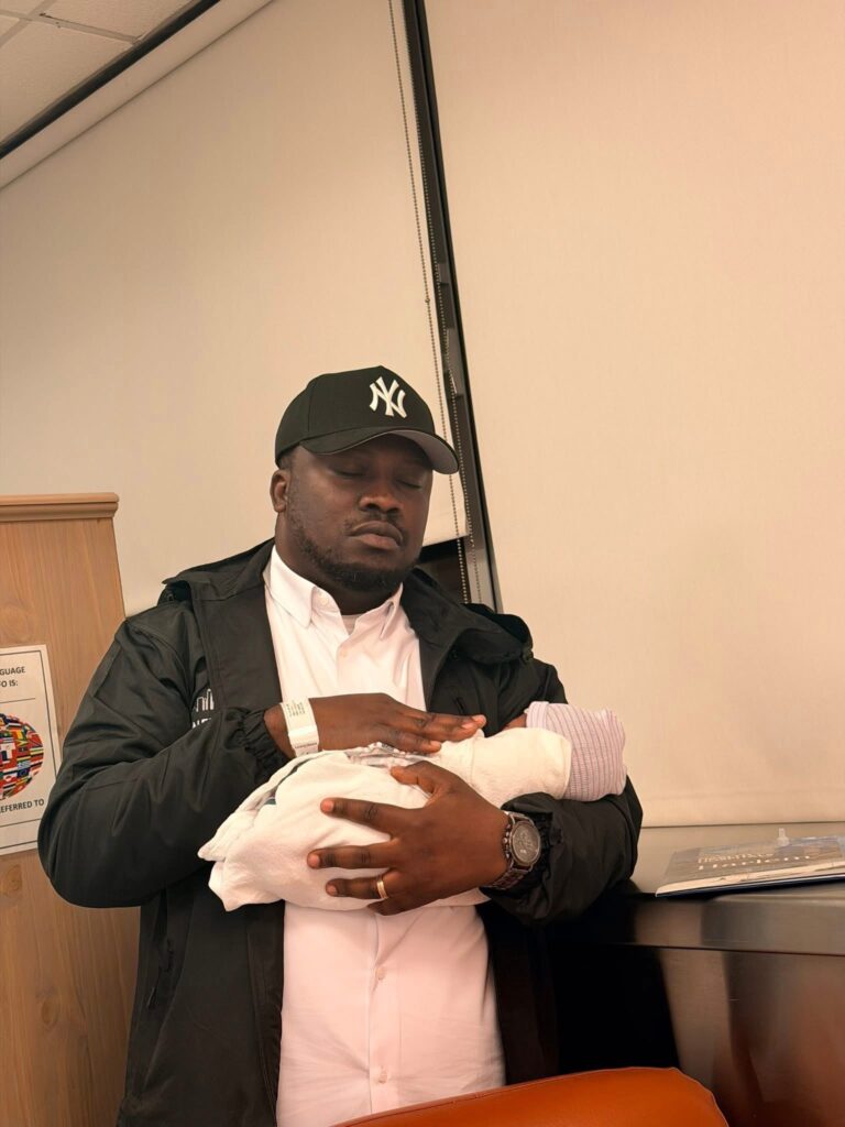 Man wearing a NY Yankees cap tenderly holding a newborn swaddled in a white blanket in a clinic setting.