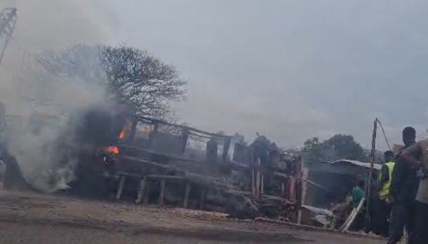 Fire and smoke rise from a collapsed wooden structure as bystanders watch outdoors.