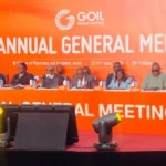 Panel of dignitaries seated at a long orange table on stage during GOIL 57th Annual General Meeting, with a bright orange backdrop and flower arrangement.