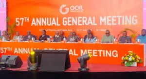 Panel of dignitaries seated at a long orange table on stage during GOIL 57th Annual General Meeting, with a bright orange backdrop and flower arrangement.