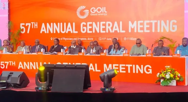 Panel of dignitaries seated at a long orange table on stage during GOIL 57th Annual General Meeting, with a bright orange backdrop and flower arrangement.