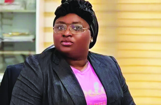 Professional woman wearing a black blazer and pink shirt with a headwrap and glasses, seated in an office.