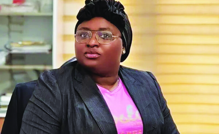 Professional woman wearing a black blazer and pink shirt with a headwrap and glasses, seated in an office.