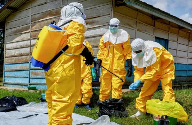 Team in full hazmat suits spray disinfectant and collect waste bags outdoors near a wooden building.
