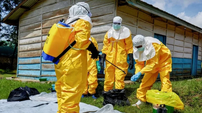 Team in full hazmat suits spray disinfectant and collect waste bags outdoors near a wooden building.