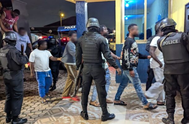 Group of civilians escorted by heavily armed police officers outside a building at night.