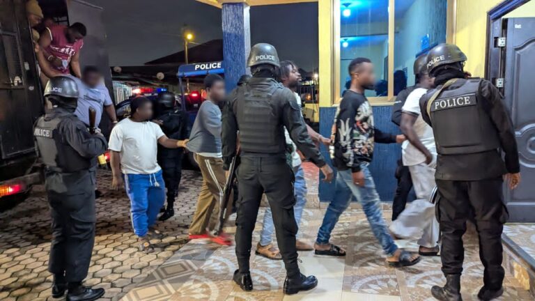 Group of civilians escorted by heavily armed police officers outside a building at night.