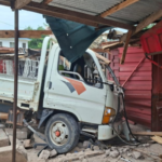 White flatbed truck crashed into a pink wooden storefront, rubble and debris scattered around while onlookers watch nearby.