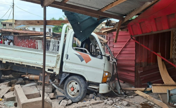 Ho-accident | Ghanaguardian.com White flatbed truck crashed into a pink wooden storefront, rubble and debris scattered around while onlookers watch nearby.