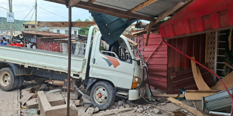 White flatbed truck crashed into a pink wooden storefront, rubble and debris scattered around while onlookers watch nearby.