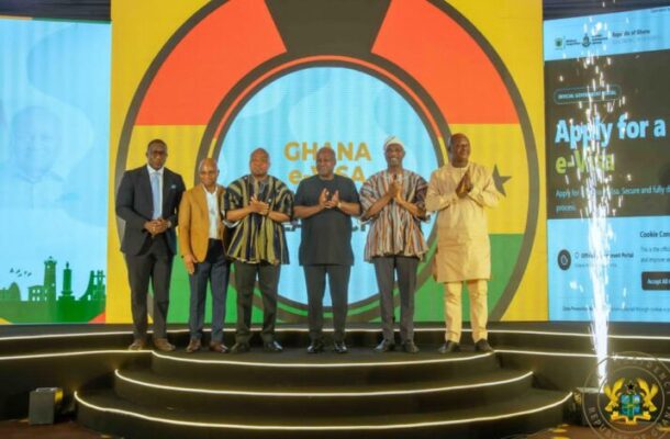 Six men in suits and traditional attire pose on a circular illuminated stage at a Ghana e-visa event, with a vibrant backdrop featuring a large round emblem and the word 'Ghana'.
