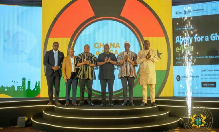 Six men in suits and traditional attire pose on a circular illuminated stage at a Ghana e-visa event, with a vibrant backdrop featuring a large round emblem and the word 'Ghana'.