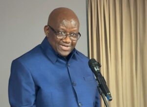 Smiling bald man wearing glasses and a blue suit speaks into a microphone indoors, with beige curtains in the background.