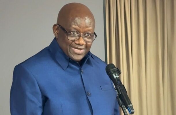 Smiling bald man wearing glasses and a blue suit speaks into a microphone indoors, with beige curtains in the background.