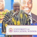 Man in striped traditional attire giving a speech at a podium labeled 'University of Education, Winneba' with microphones in front of him.