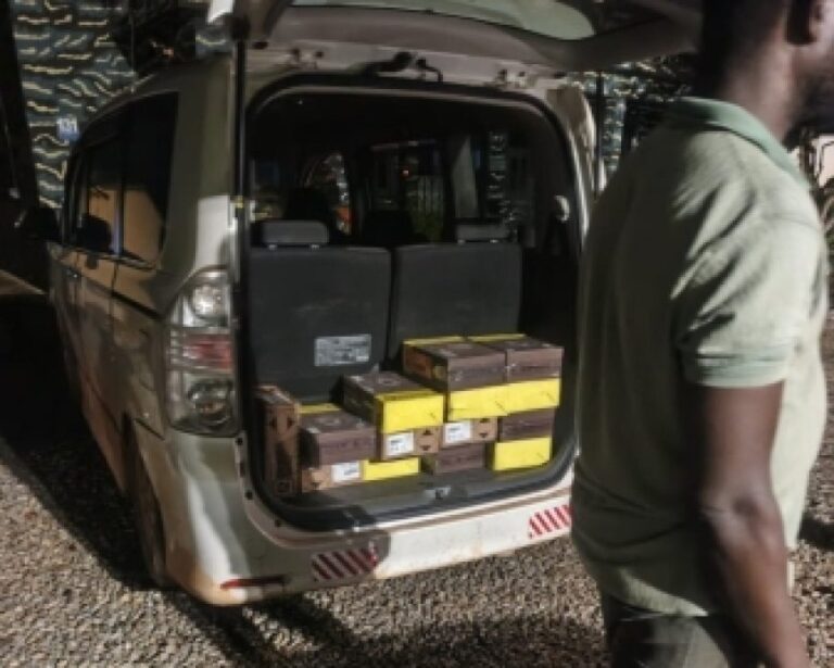 Open rear of a beige minivan loaded with stacked yellow and brown boxes; a man wearing a green polo stands to the right.