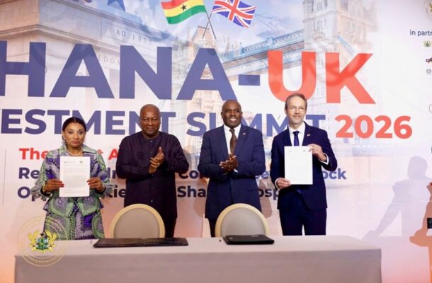 Four officials on stage at the Ghana-UK Investment Summit 2026, two holding signed documents as others applaud, flags in the background.
