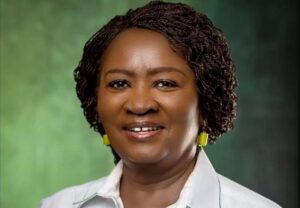 Portrait of a smiling Black woman with short braided hair, wearing a white collared shirt and yellow earrings, against a green studio backdrop.