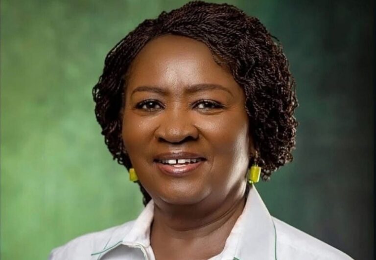 Portrait of a smiling Black woman with short braided hair, wearing a white collared shirt and yellow earrings, against a green studio backdrop.