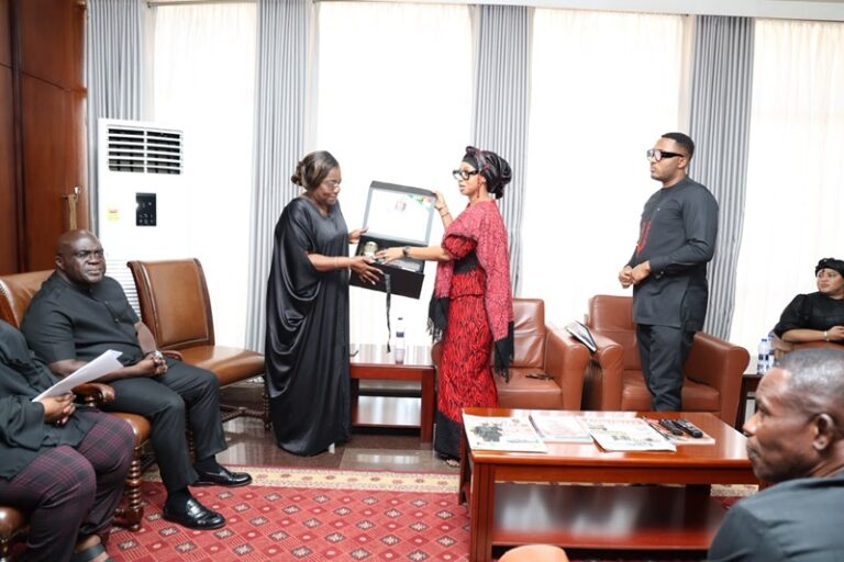 Two women exchange a framed item in a formal living room as several people watch, seated on leather chairs and sofas around a coffee table.