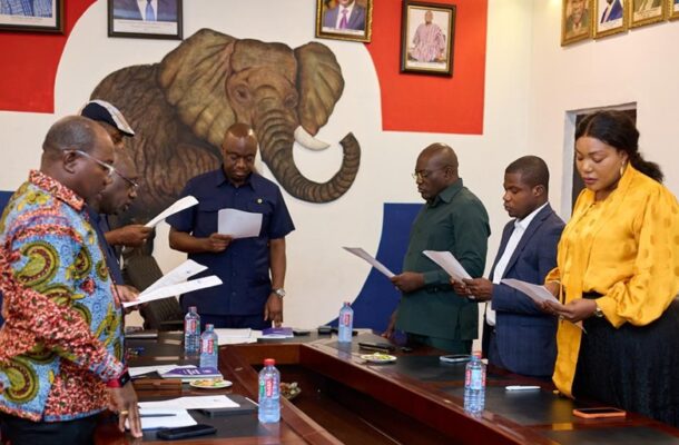 Six professionals stand around a conference table, reviewing documents, with a large elephant mural on the wall behind them.