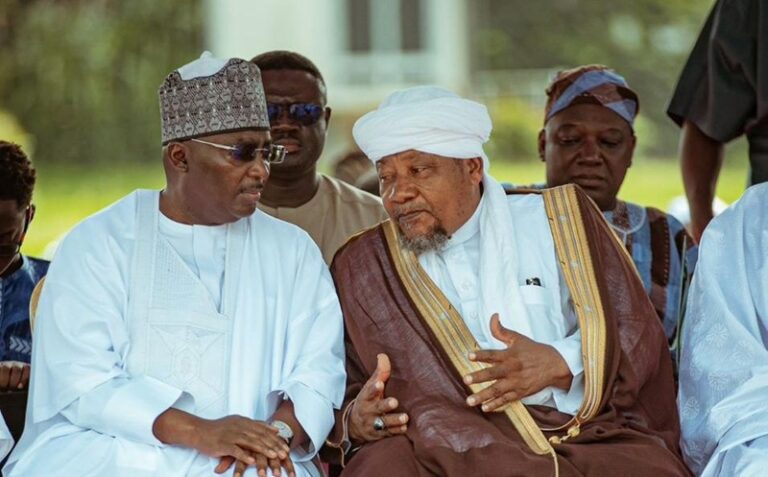 Two men in traditional white and brown robes sit outdoors, talking in conversation as others look on.