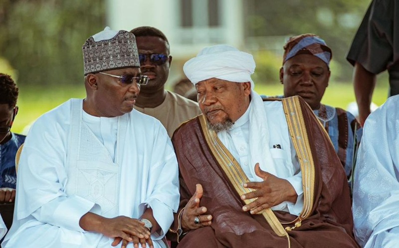 Two men in traditional white and brown robes sit outdoors, talking in conversation as others look on.